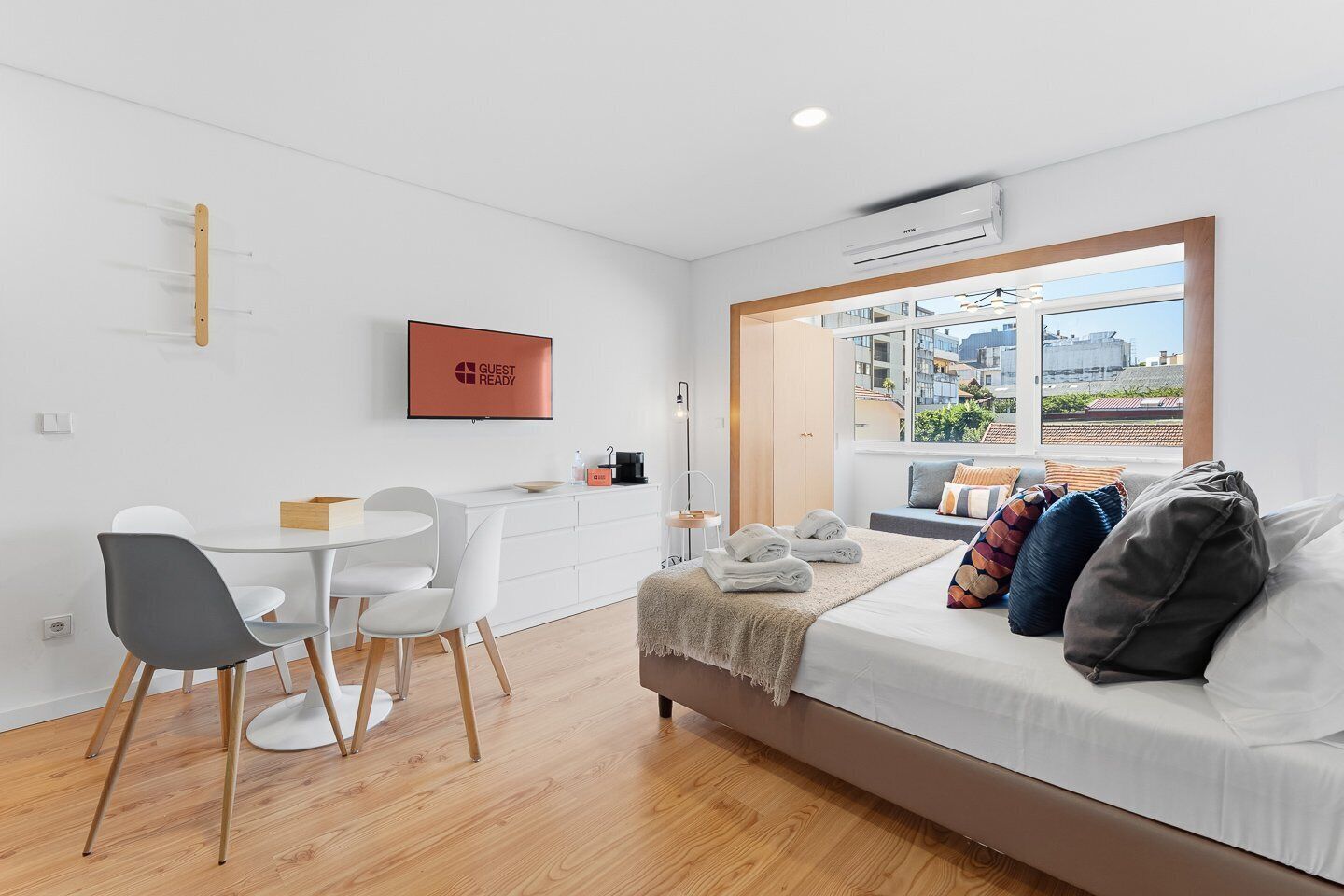 A modern, well-lit room with a dining area, a sofa with cushions, a TV, and a window with a city view.