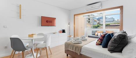 A modern, well-lit room with a dining area, a sofa with cushions, a TV, and a window with a city view.
