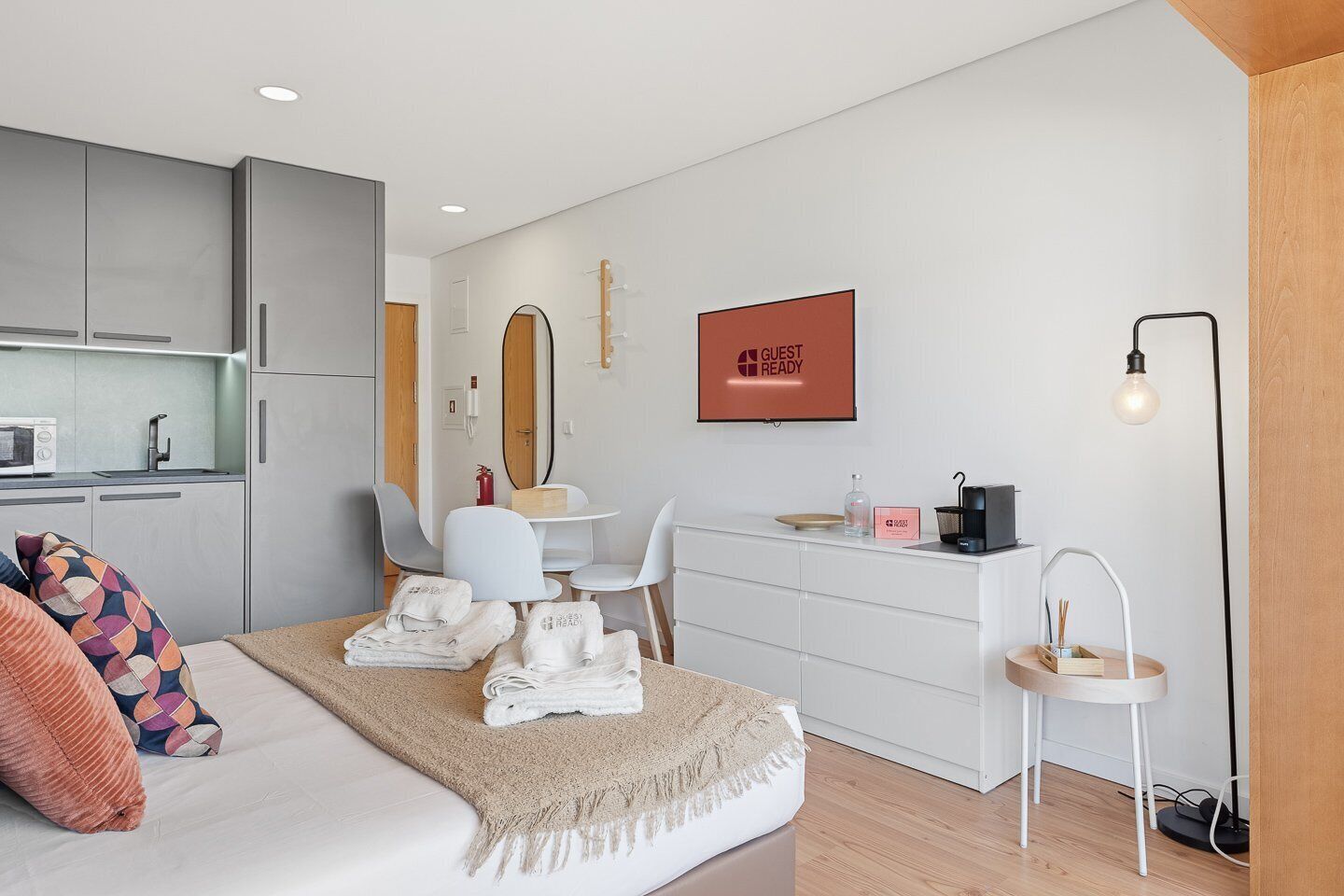 A modern hotel room with a bed, a desk, a chair, a small table, a mirror, a TV, and a bedside lamp.