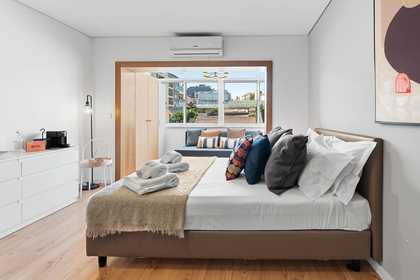 A modern bedroom with a bed, a view of the city, and a kitchenette area.