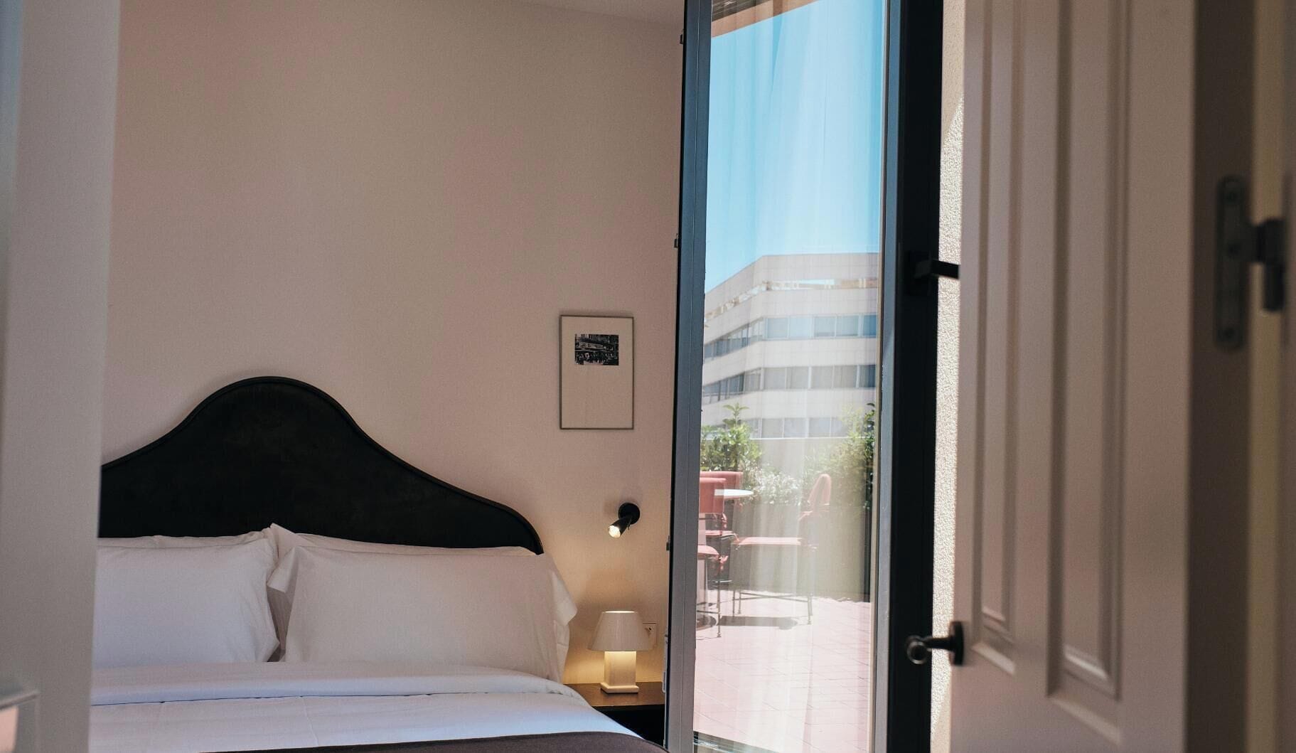 Une chambre d’hôtel avec un lit, une table de chevet avec une lampe, un tableau encadré au mur et une vue sur l’extérieur.