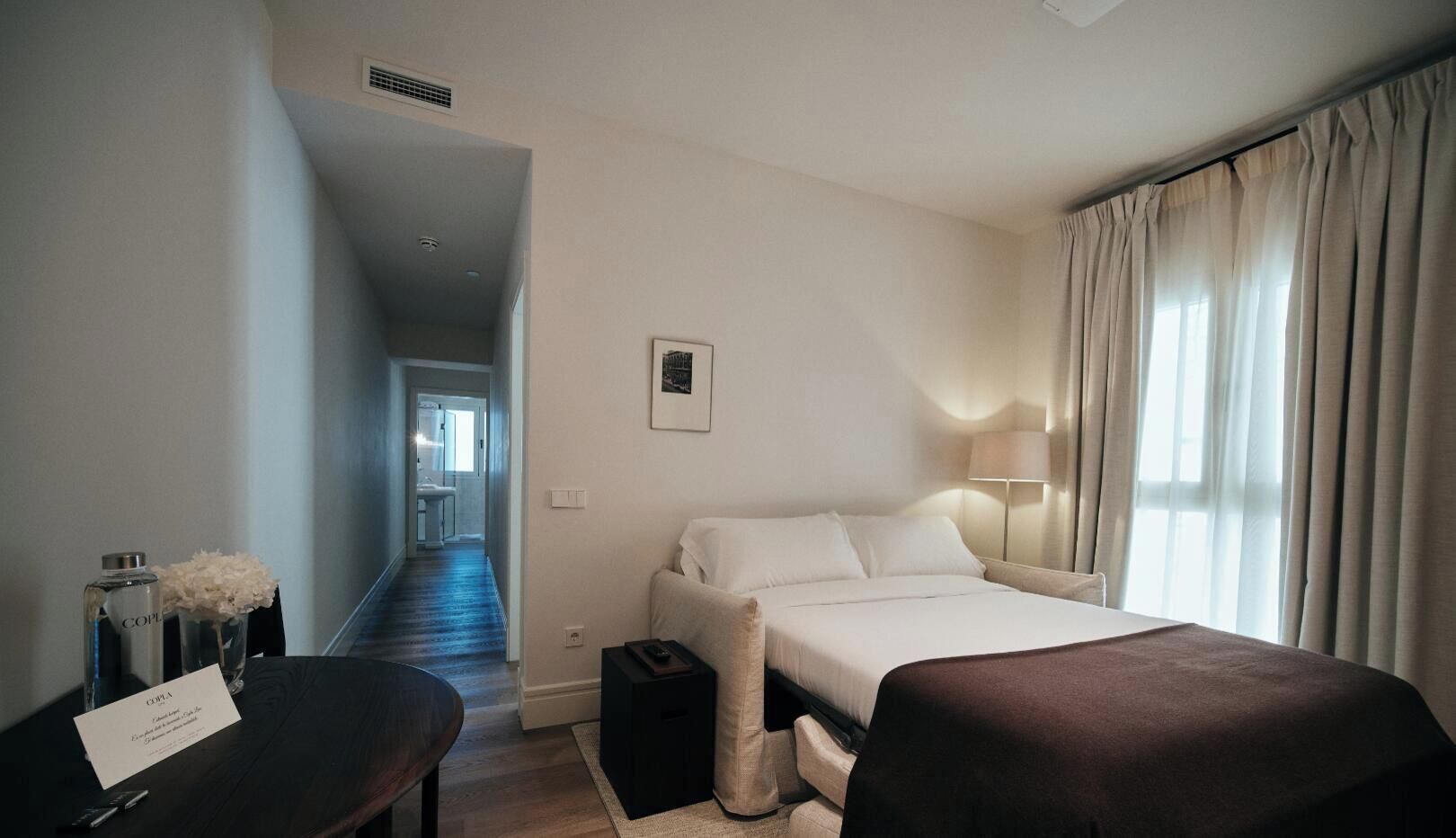 A hotel room with a bed, bedside table, lamp, and a view of a corridor.