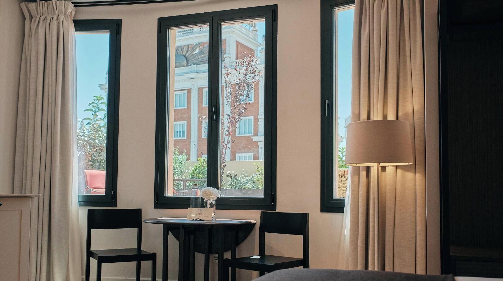 Una habitación con una mesa, sillas, una lámpara y una ventana que ofrece vistas a edificios.