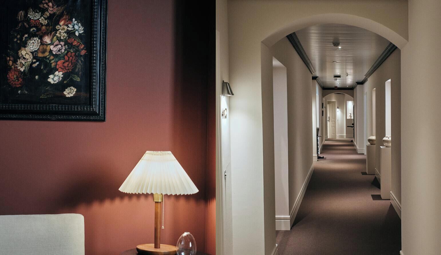 A hotel corridor with a lamp, a framed floral artwork, and a view of the hallway leading to a door.