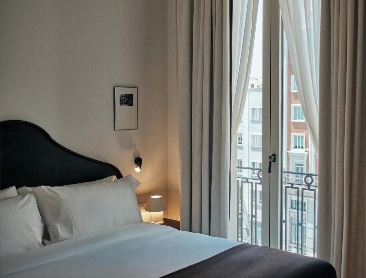 Ein Hotelzimmer mit einem Bett, einem Nachttisch, einer Lampe und Blick auf Gebäude durch ein Fenster.
