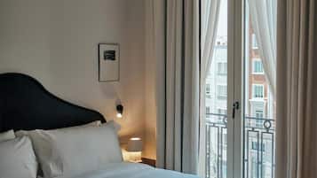 A hotel room with a bed, a nightstand, a lamp, and a view of buildings through a window.