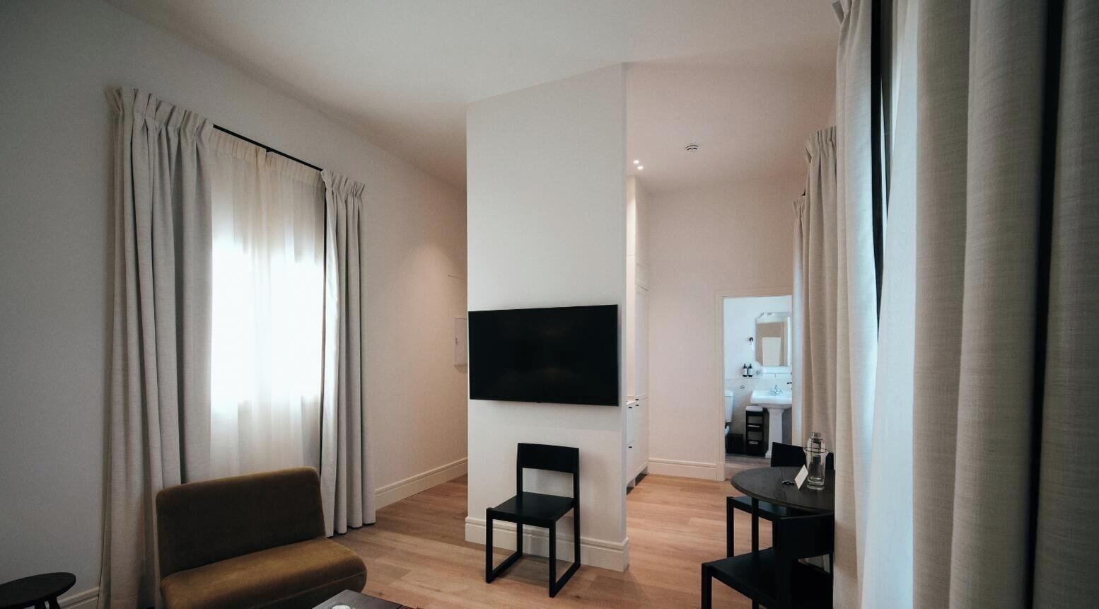 Habitación de hotel con una televisión de pantalla plana montada en la pared, un sofá, una mesa de comedor con sillas y una cocina al fondo.