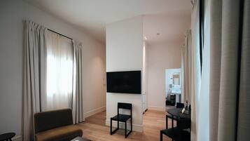 Habitación de hotel con una televisión de pantalla plana montada en la pared, un sofá, una mesa de comedor con sillas y una cocina al fondo.