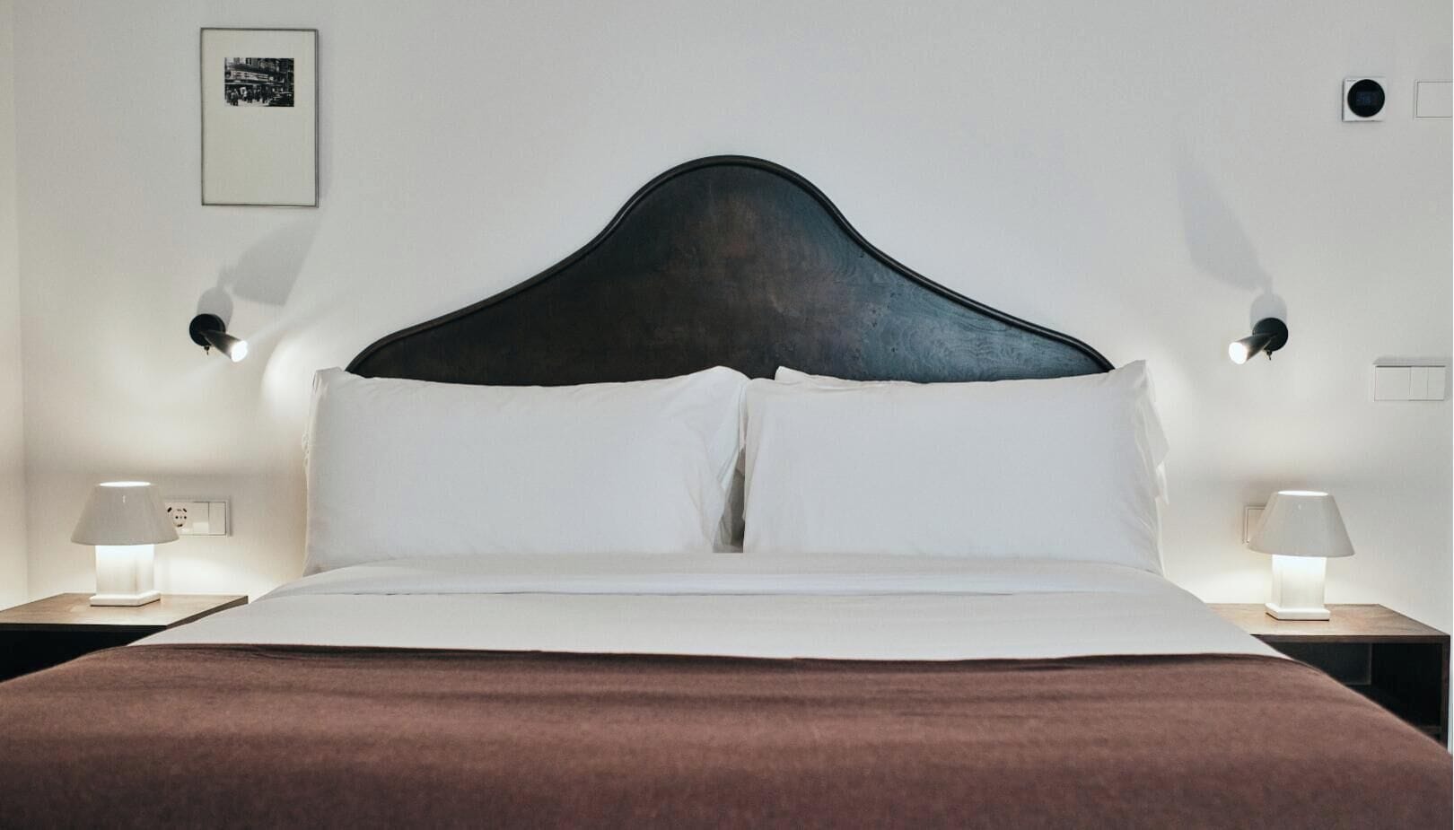 A hotel room with a large bed, two bedside tables with lamps, a framed picture on the wall, and a thermostat.