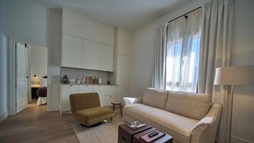 Ein modernes Wohnzimmer mit einer Couch, einem Sessel und einem Couchtisch. Es gibt eine Küchenzeile mit Schränken und einem Esstisch.