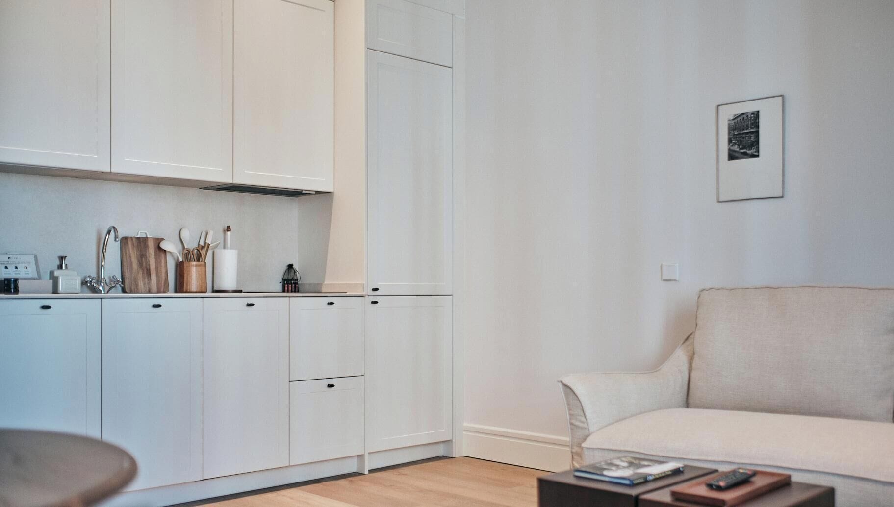Una cocina moderna con armarios blancos y un horno empotrado, un sofá color beige y una pequeña mesa con libros y un control remoto.