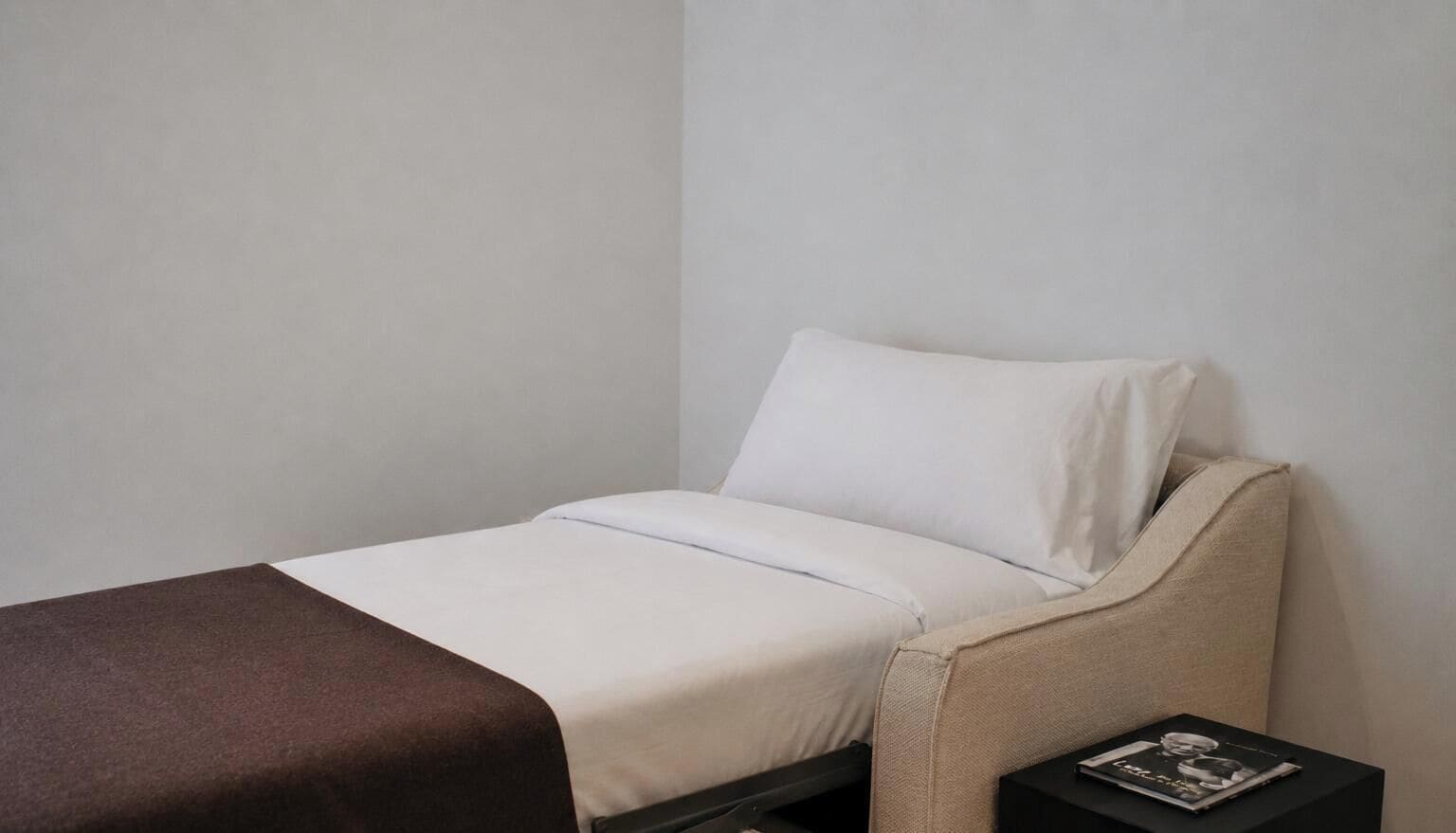A single bed with a white mattress and a brown blanket, a beige upholstered headboard, and a small black nightstand with a magazine.