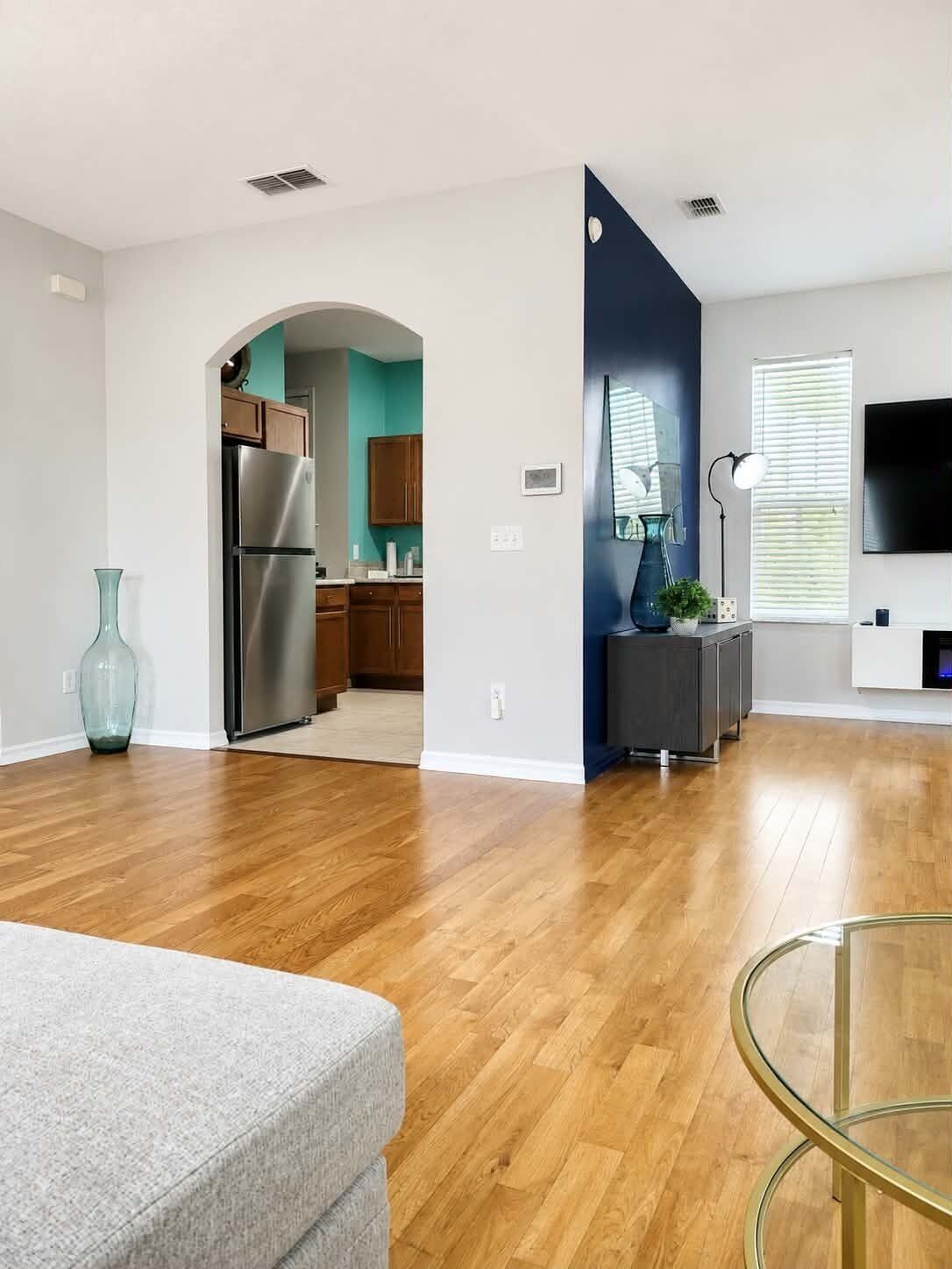 A modern living room with hardwood flooring, a grey sofa, a glass coffee table, and a kitchen area with wooden cabinets and a stainless steel refrigerator.