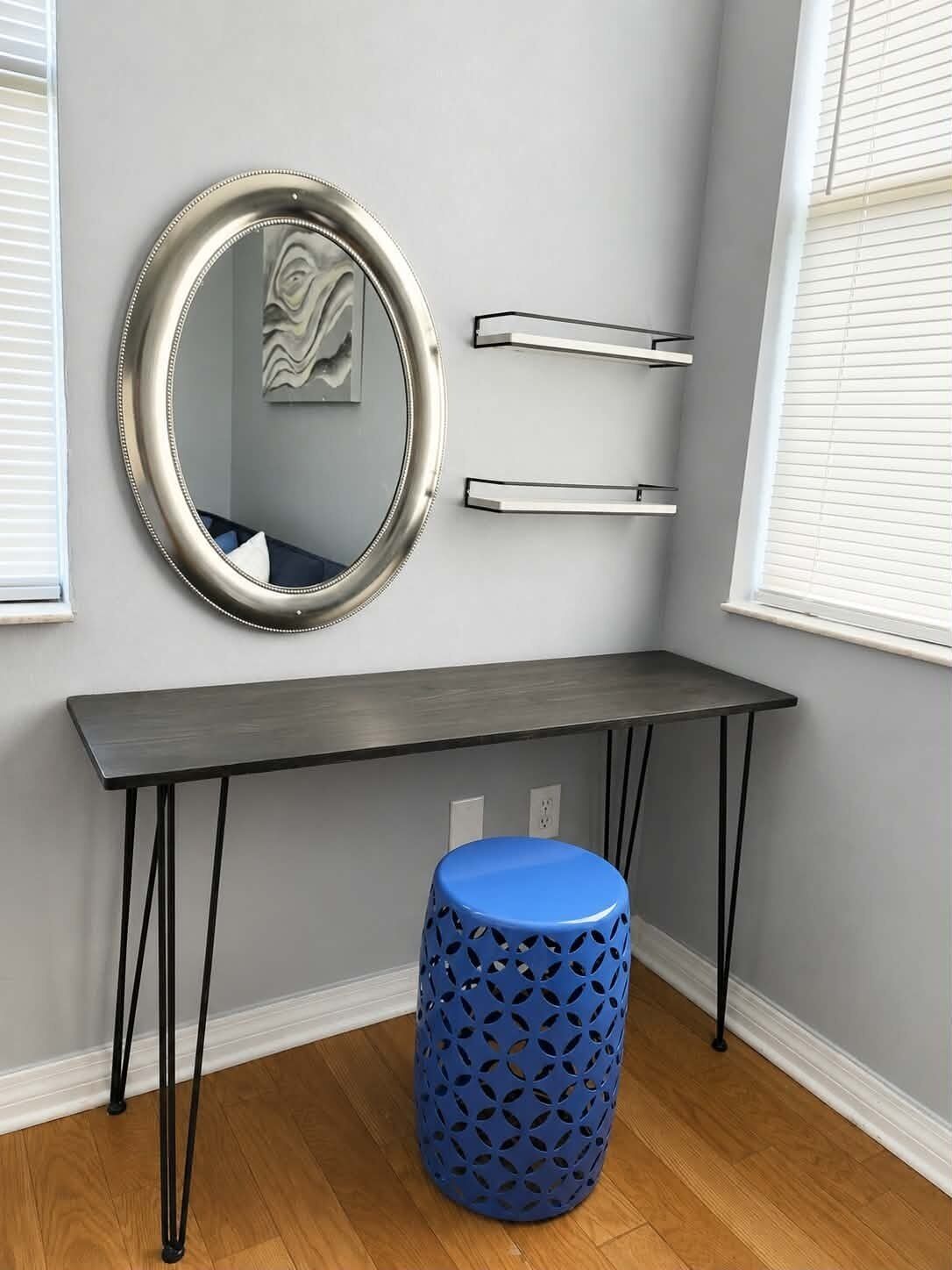 A room with a grey wall, a round mirror, a black table with metal legs, and a blue stool with a geometric pattern.