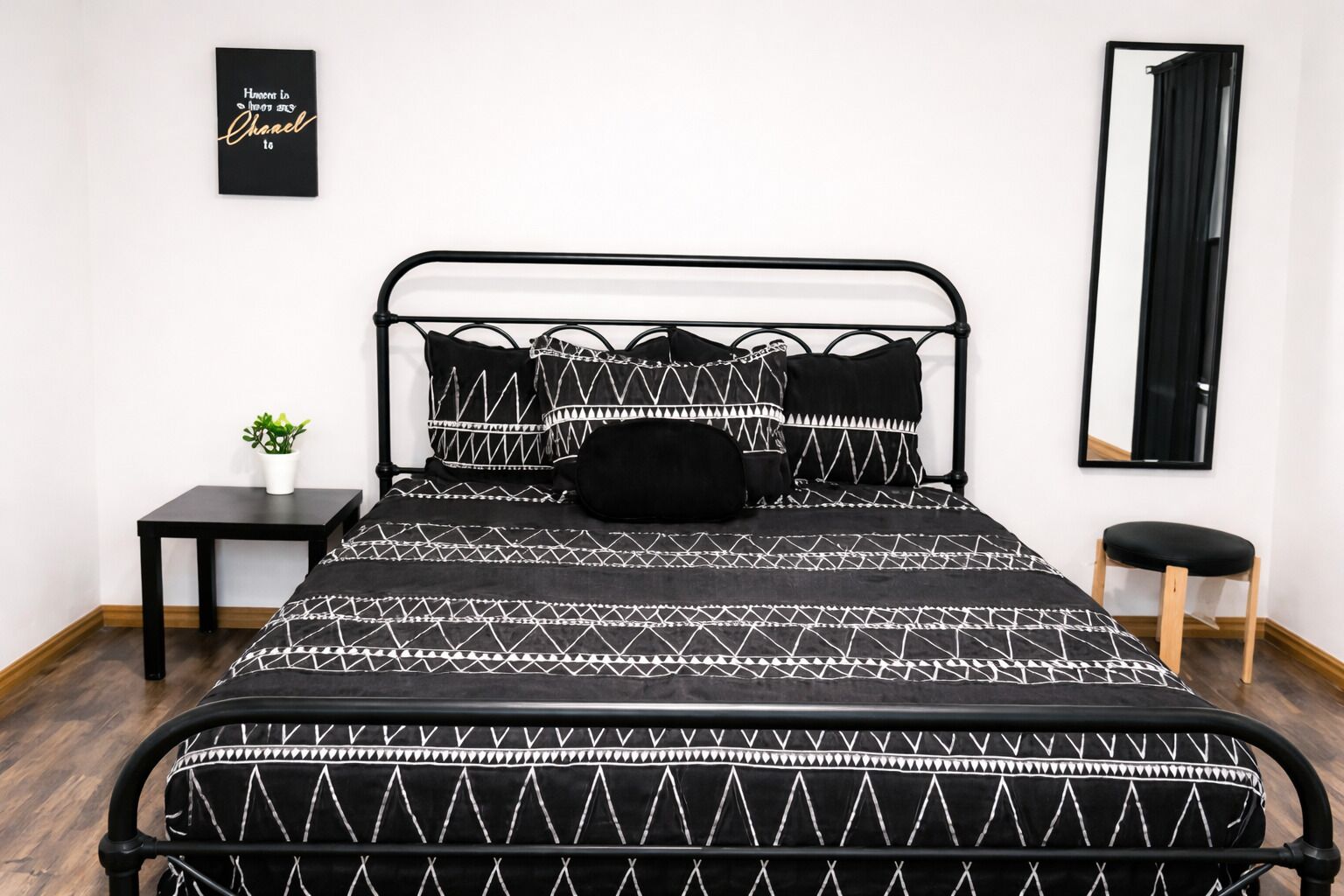 Un lit avec un cadre en métal noir, une literie à motifs noir et blanc, une petite table de chevet avec une plante et un miroir accroché au mur.