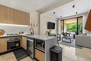 A modern kitchen with a central island, a dining area with a sofa and coffee table, and a large sliding glass door leading to an outdoor space. - Yáax K'iin: Poolside Oasis + Free E-Bikes (Playa del Carmen)