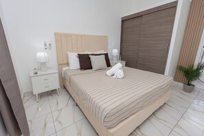 A hotel room with a large bed, a nightstand with a lamp, a potted plant, and a sliding door. - Modern 1BR in Plaza Clara in Bayahibe (San Rafael del Yuma)