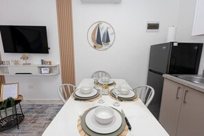 A dining area with a white table set for four, a television mounted on the wall, and a refrigerator in the kitchen area. - Modern 1BR in Plaza Clara in Bayahibe (San Rafael del Yuma)