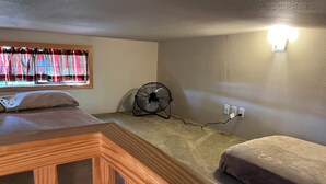 A bedroom with a wooden bed frame, a window with curtains, a standing fan, and a wall-mounted light fixture.