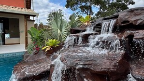A pool with a waterfall feature, surrounded by tropical plants and a building with a tiled roof. - OKA VIVAH - Chalet Oka das Aguas (Jaboticatubas)
