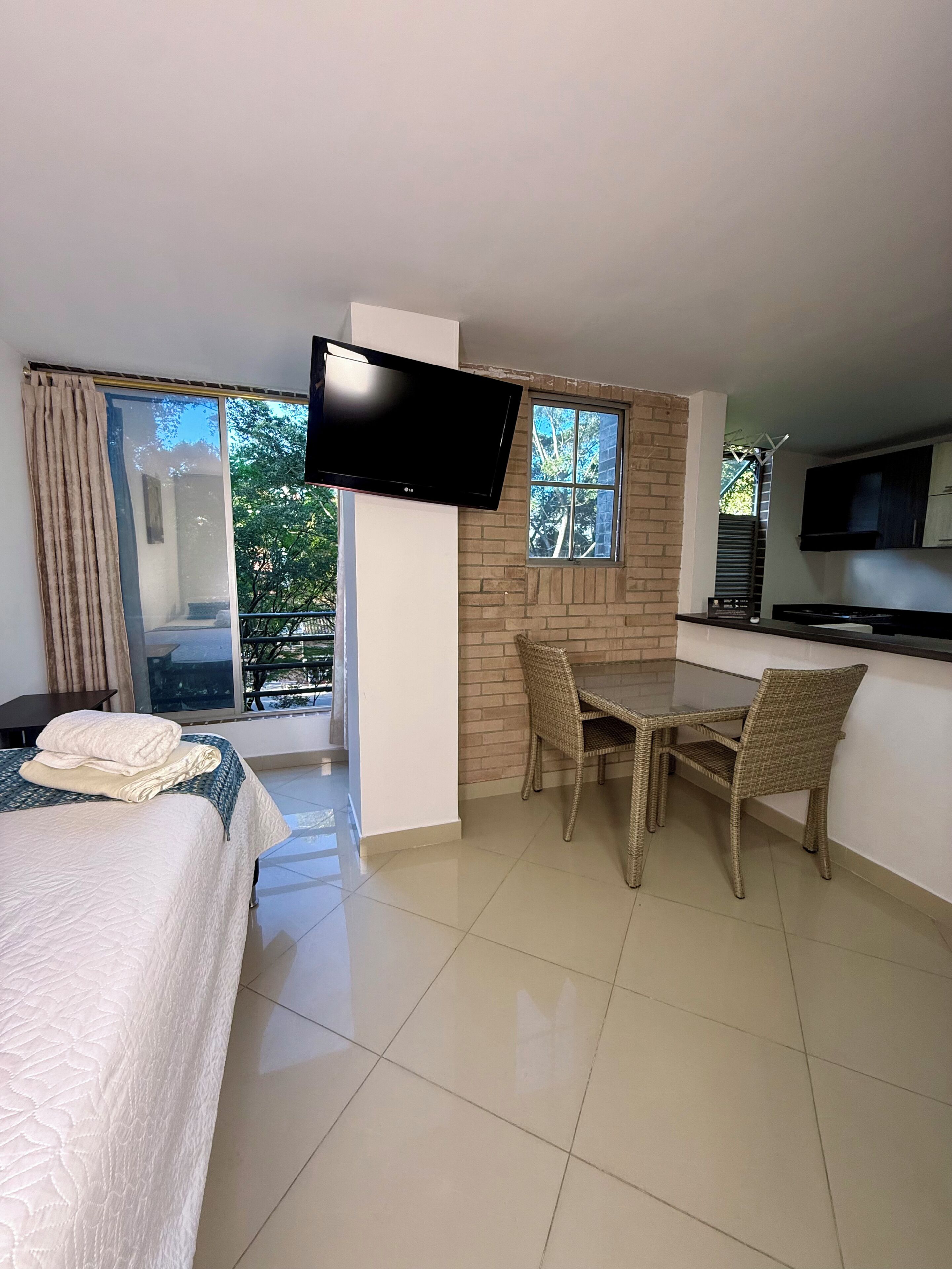 A hotel room with a bed, a dining table with chairs, a flat-screen TV mounted on the wall, and a window with a view of trees.