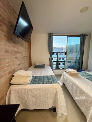 A hotel room with two beds, a flat-screen TV mounted on the wall, and a balcony with a view of buildings. - Living Montes (MedellÃn)