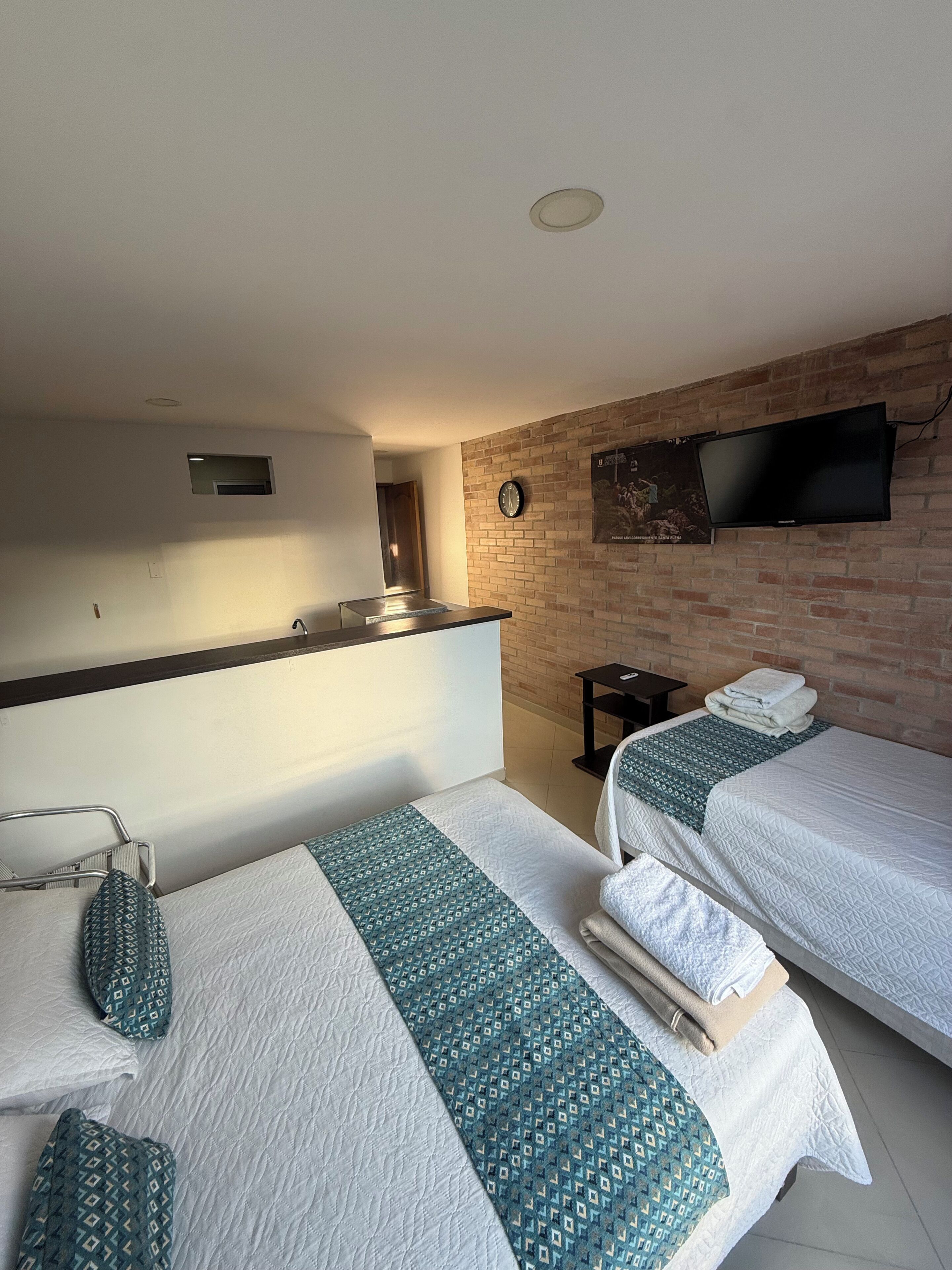 A hotel room with two beds, a TV mounted on a brick wall, a small table, and a clock.