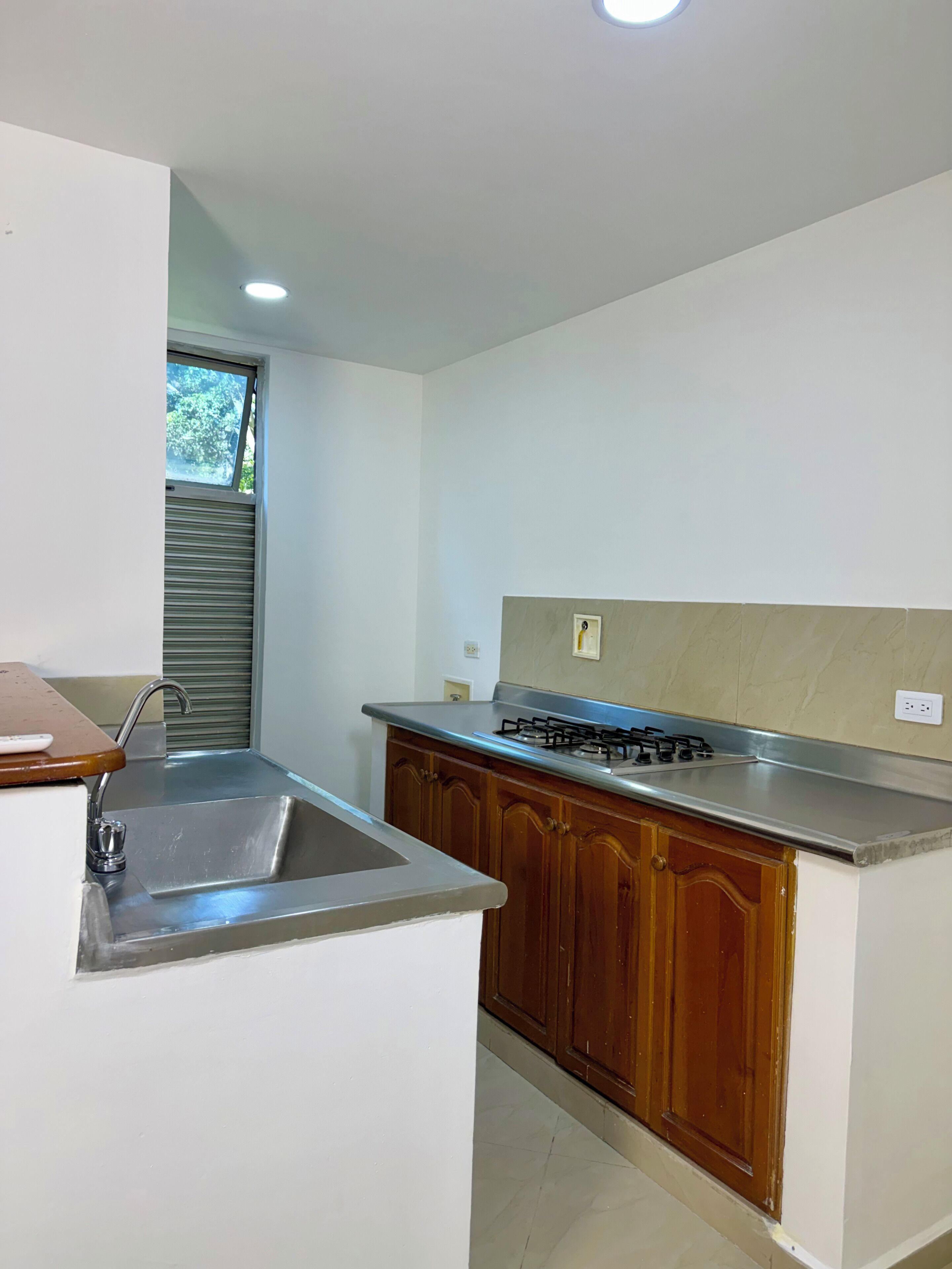 Una cocina con un lavamanos de acero inoxidable, armarios de madera, una estufa a gas y una ventana con vista a vegetación.