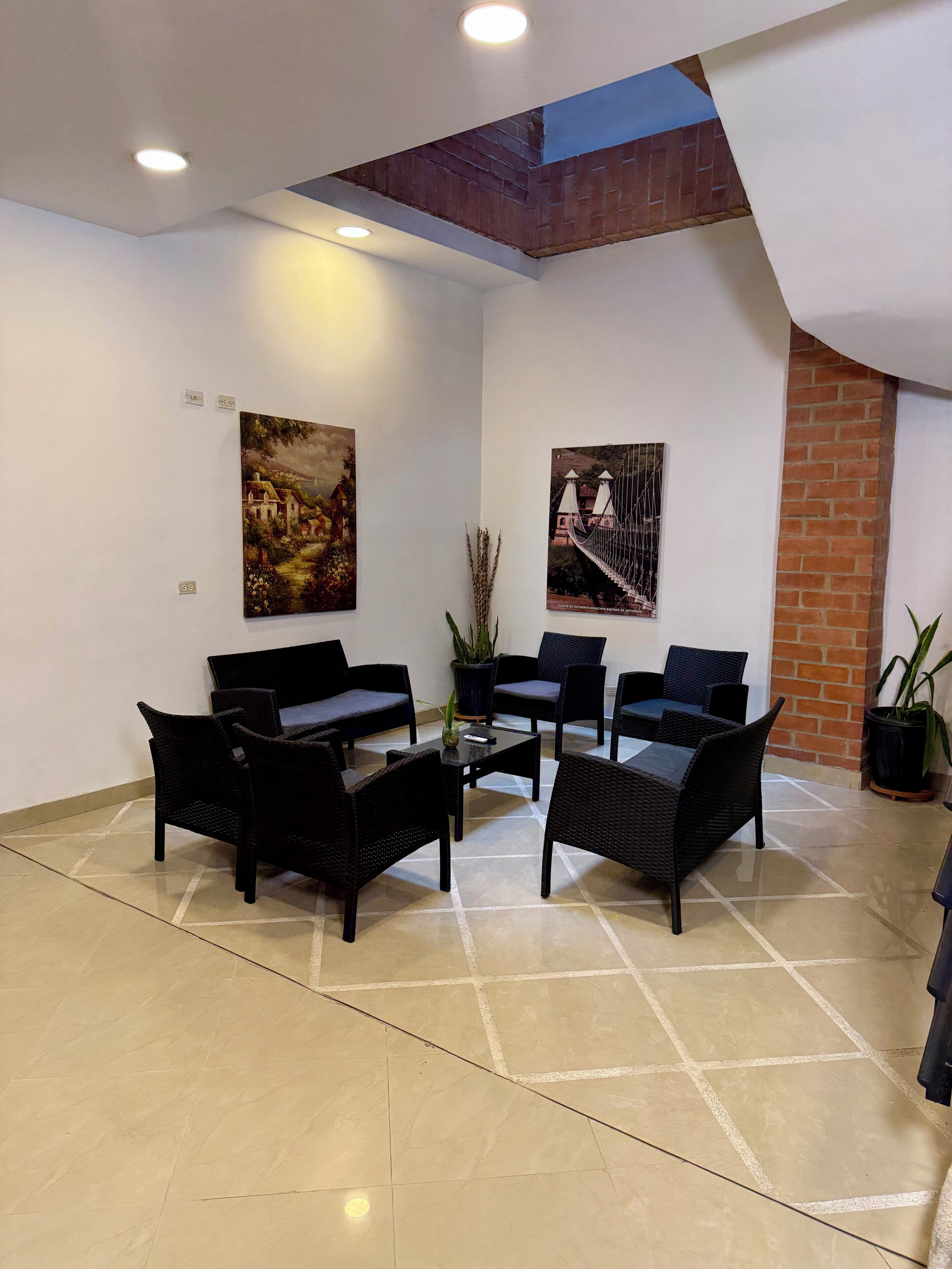 Una habitación con piso de baldosas, mobiliario de mimbre negro, dos cuadros en la pared y una pared de ladrillo.