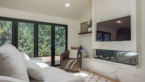 A modern living room with a sofa, a fireplace, and a large window with a view of greenery. - Luxury Capitola Retreat | Hot Tub | Blocks to Sand (Capitola)