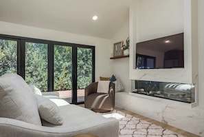 A modern living room with a sofa, a fireplace, and a large window with a view of greenery. - Luxury Capitola Retreat | Hot Tub | Blocks to Sand (Capitola)