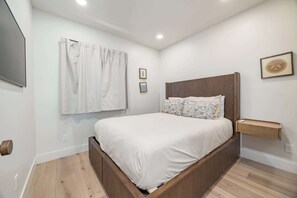 A modern bedroom with a wooden bed frame, white bedding, a wall-mounted TV, and a bedside table with a phone. - Luxury Capitola Retreat | Hot Tub | Blocks to Sand (Capitola)