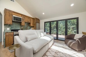 A modern living space with a sofa, a coffee table, a chair, a kitchen area with wooden cabinets, and a large window offering a view of greenery. - Luxury Capitola Retreat | Hot Tub | Blocks to Sand (Capitola)