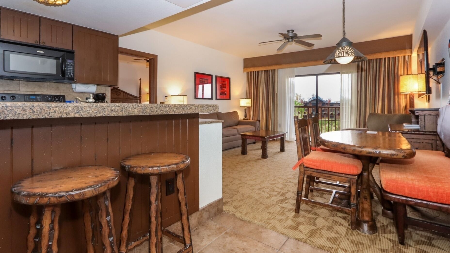 A kitchen with a microwave, wooden bar stools, a dining table with chairs, a ceiling fan, and a view of the outdoors through a window.