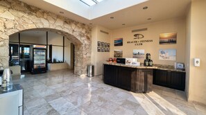 A reception area with a stone archway, a refrigerated display case, a counter with health supplements, and a wall with fitness club posters. - Beachfront King Studio at Diamante Dunes (Cabo San Lucas)