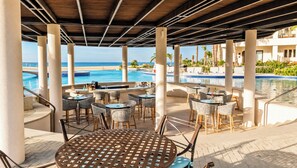 A covered outdoor seating area with tables and chairs, overlooking a pool and the ocean. - Beachfront King Studio at Diamante Dunes (Cabo San Lucas)