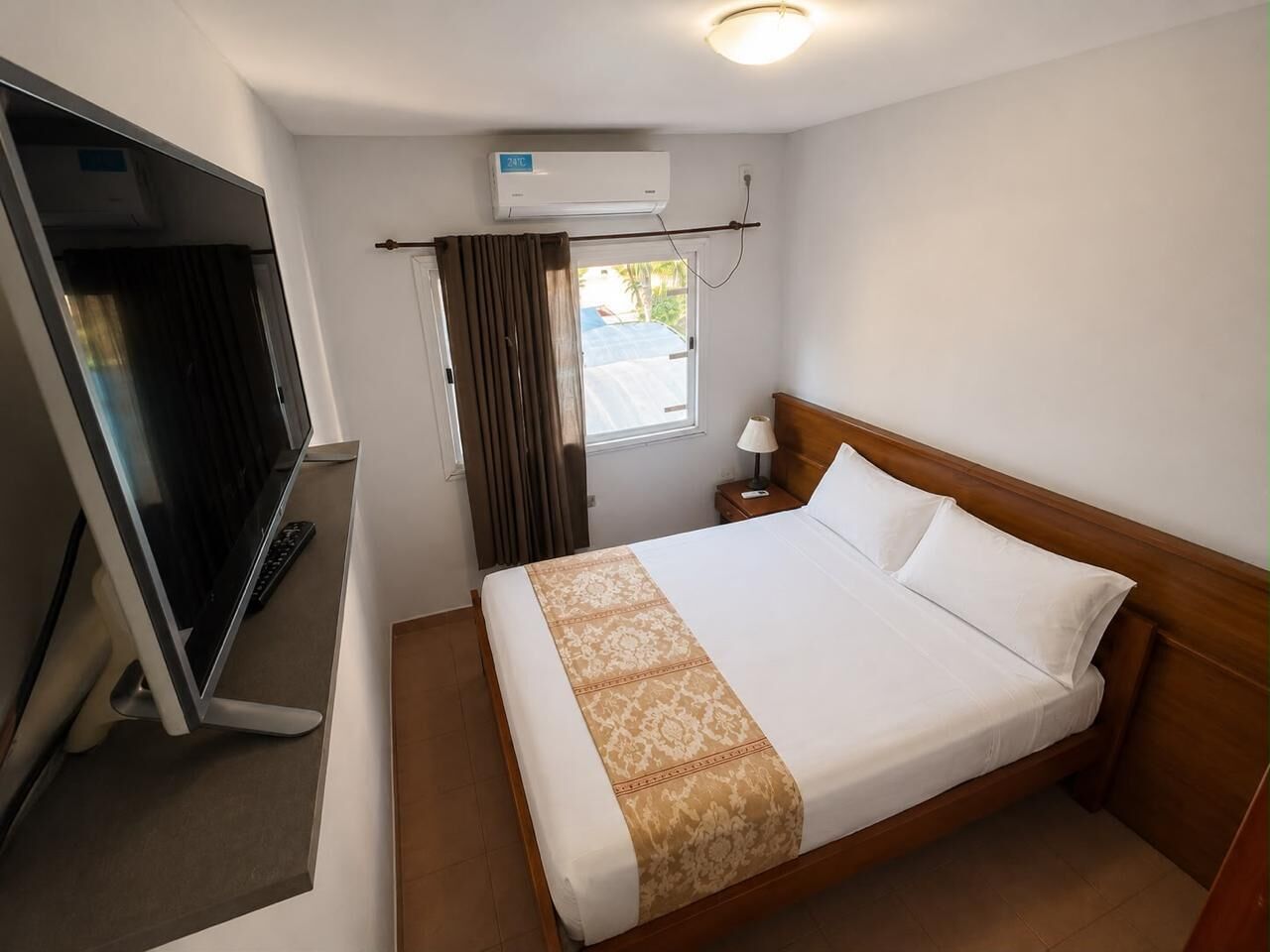 A hotel room with a bed, a TV, an air conditioning unit, and a window with curtains.