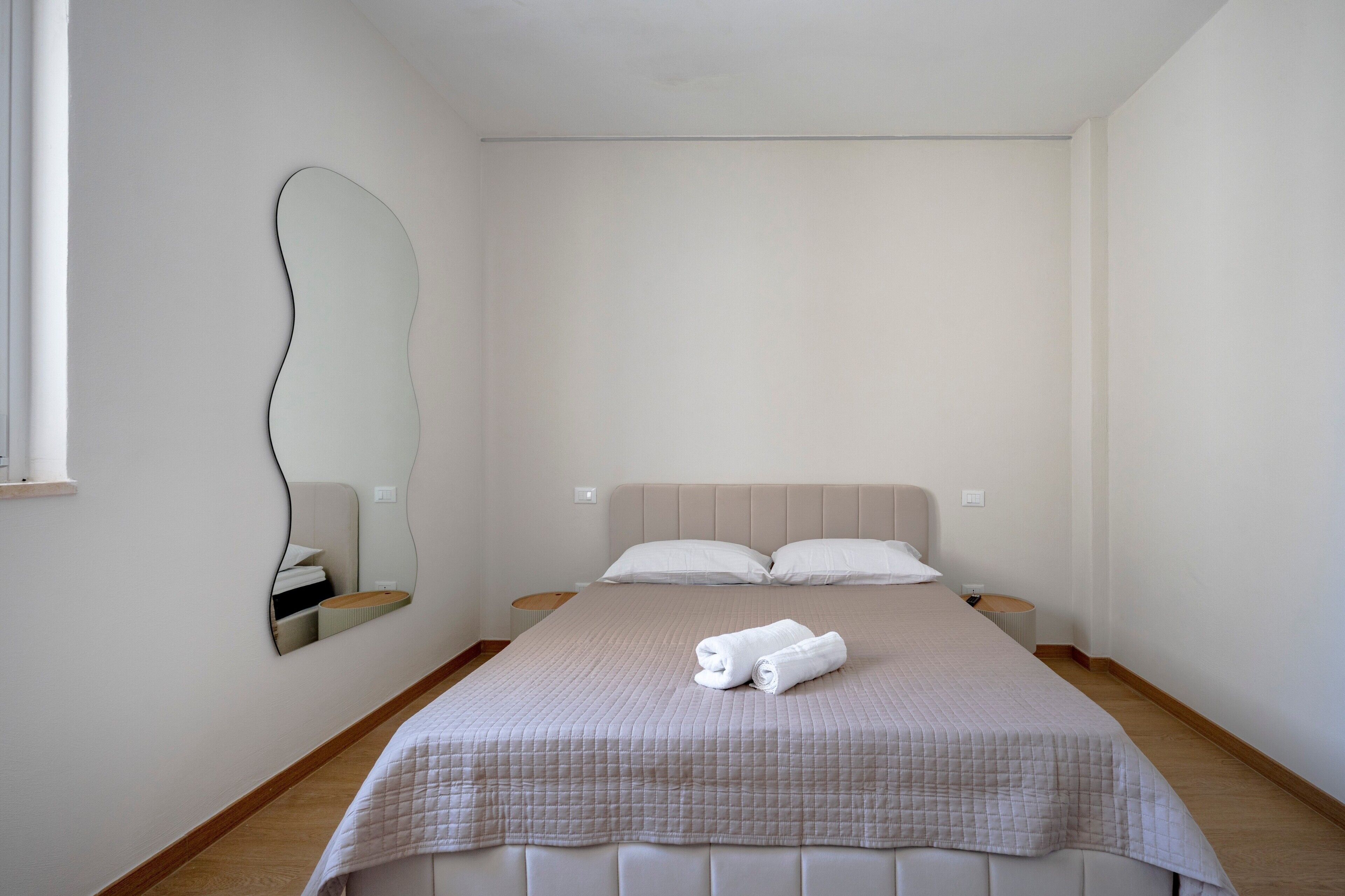 Ein schlicht eingerichtetes Schlafzimmer mit einem Bett, zwei Nachttischen, einem Spiegel und einem Fenster.