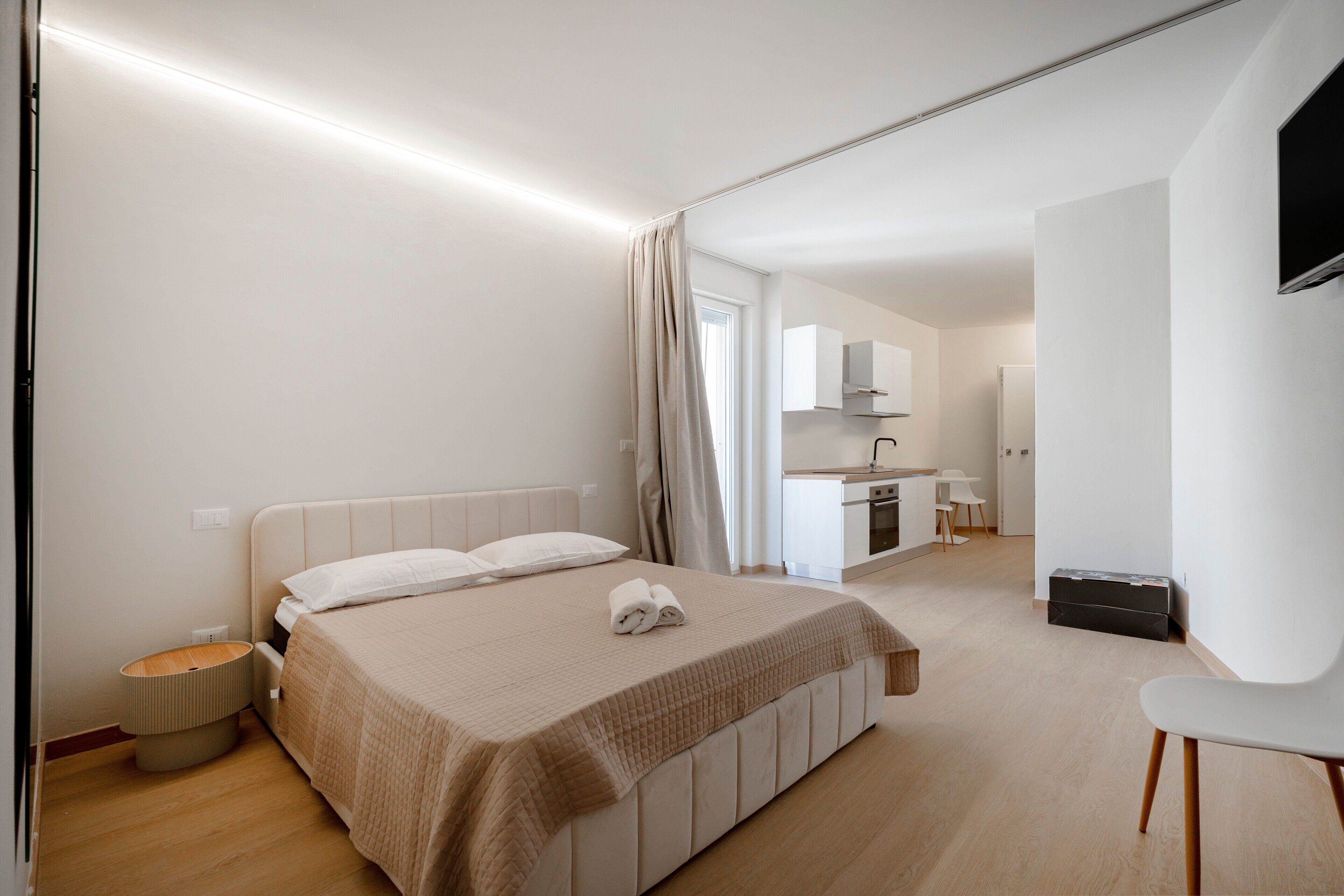 Une chambre d’hôtel moderne avec un lit, une petite table de chevet, une chaise et, au fond, une kitchenette.