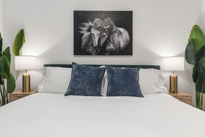 Uma cama com lençóis brancos, duas almofadas azuis e uma pintura de um cavalo preto e branco acima.