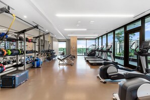 Academia moderna com equipamentos de musculação, esteiras e vista para edifícios através de amplas janelas.