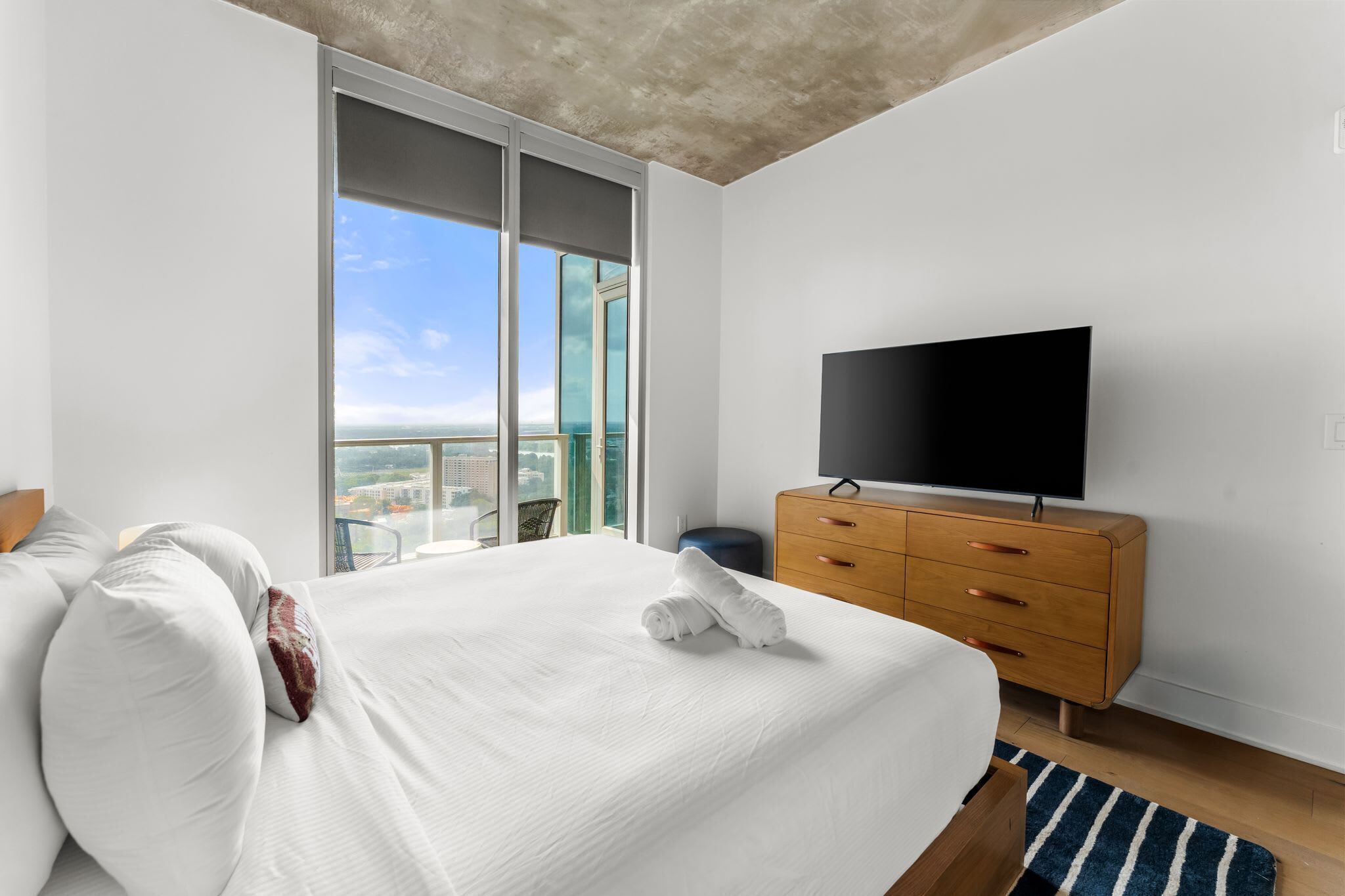 A hotel room with a large bed, a flat-screen TV, a wooden dresser, and a balcony with a city view.