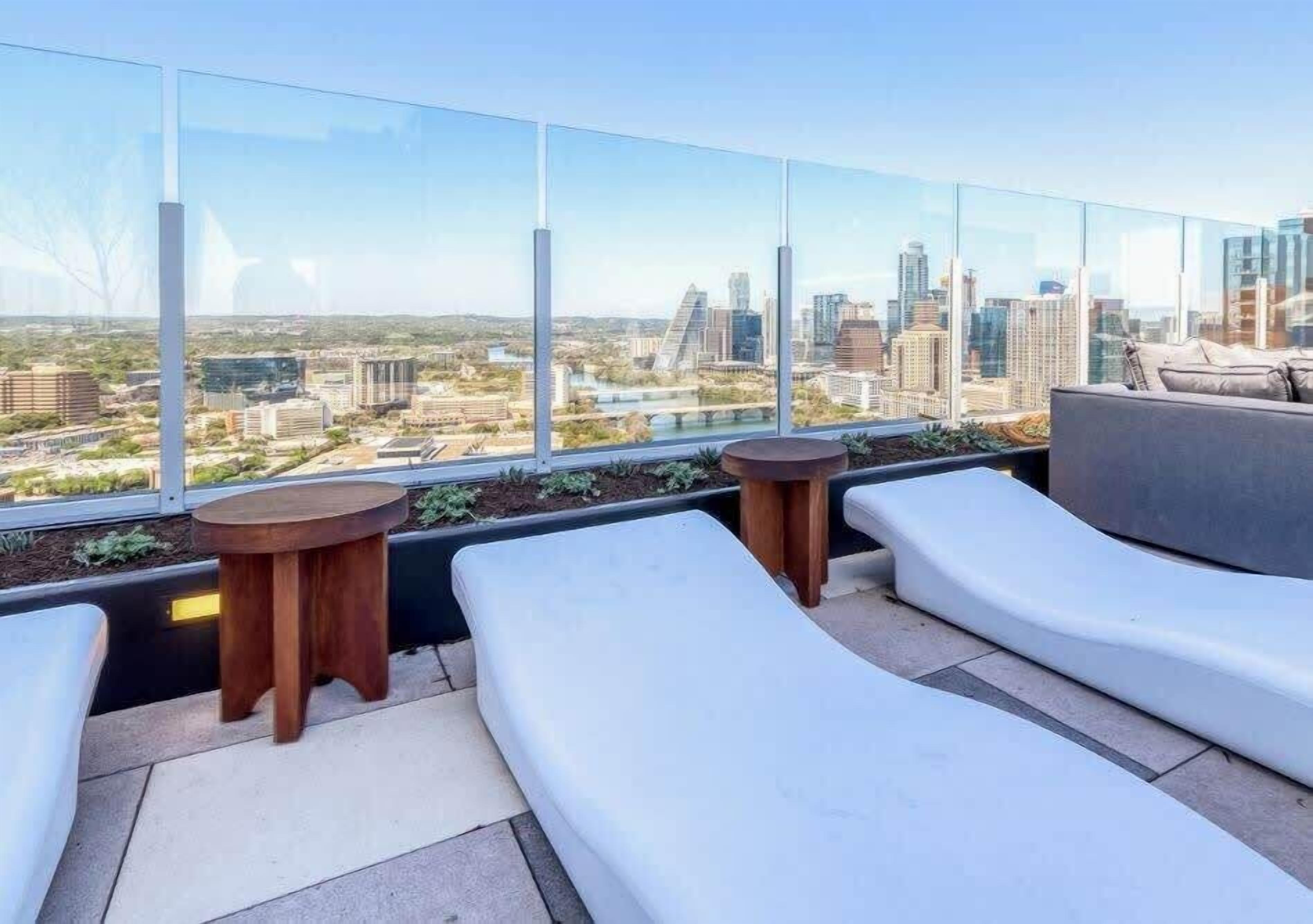 A rooftop terrace with white lounge chairs, a round wooden table, and a view of a cityscape.