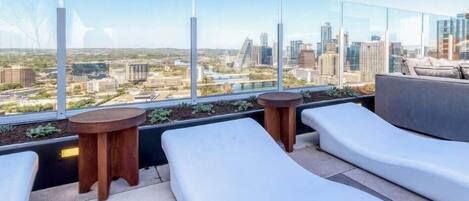 A rooftop terrace with white lounge chairs, a round wooden table, and a view of a cityscape.
