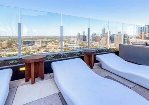 A rooftop terrace with white lounge chairs, a round wooden table, and a view of a cityscape. - Downtown Skyline & Lake Views l Rooftop Pool (Austin)