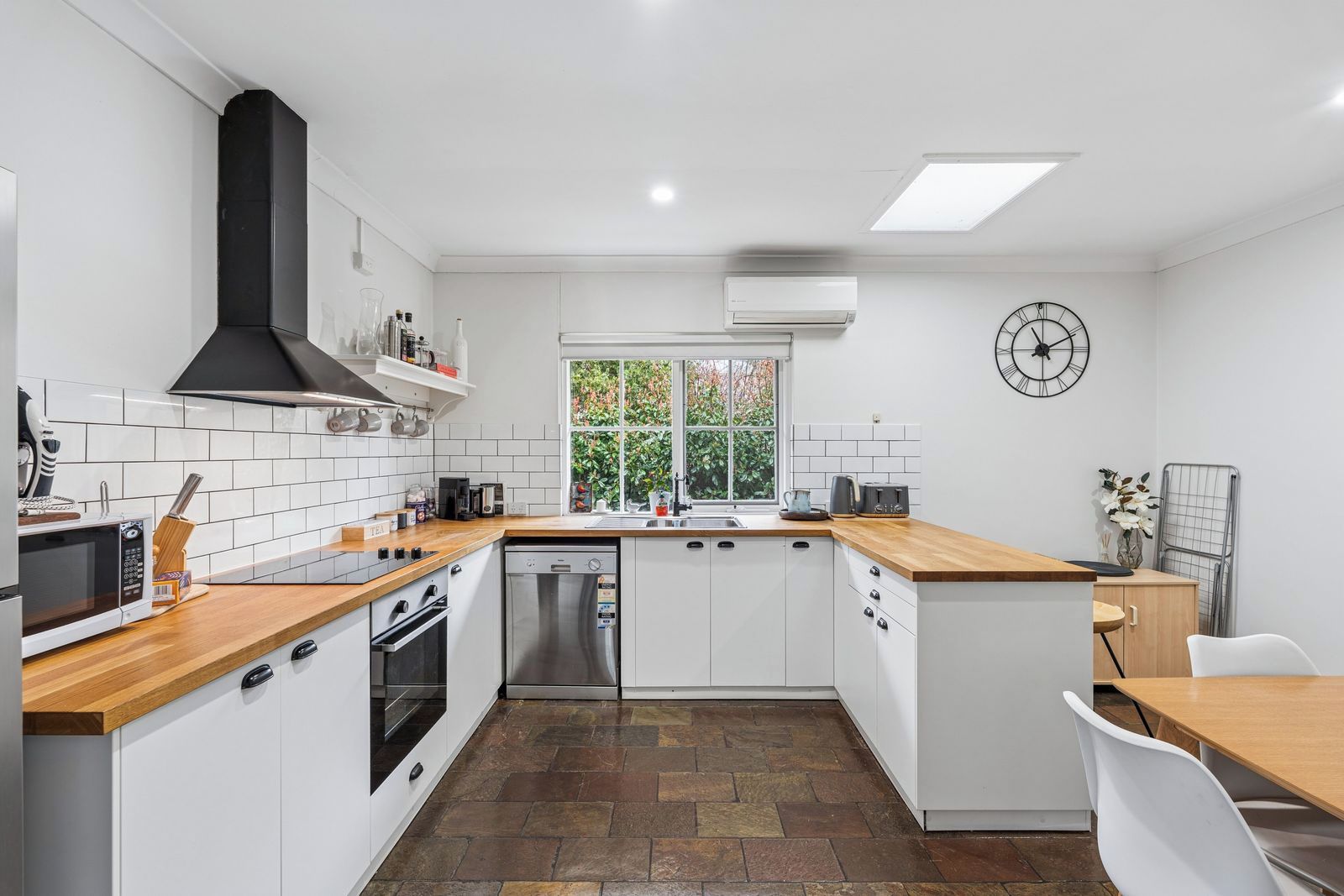 A modern kitchen with white cabinetry, a black range hood, a built-in dishwasher, and a window with a view of greenery.