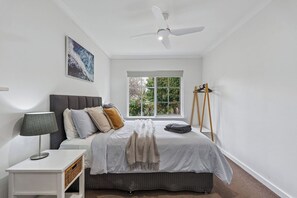 Un dormitorio con cama, mesita de noche, lámpara, ventana con vista a follaje y ventilador de techo.