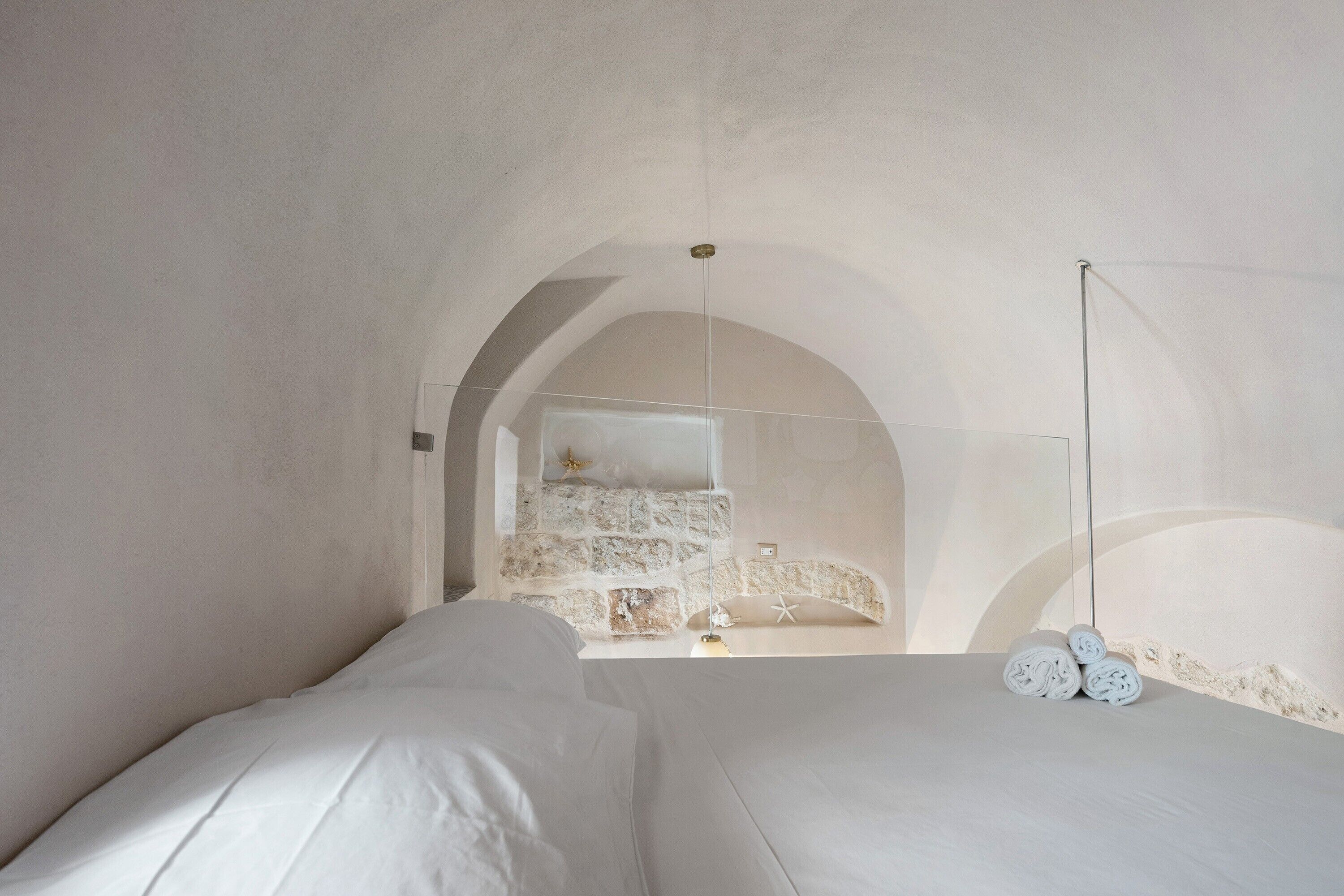 Une pièce minimaliste avec une grande porte en arc donnant sur un mur de pierre. On y trouve deux lits avec du linge de lit blanc et une paire de pantoufles blanches posées sur le sol.