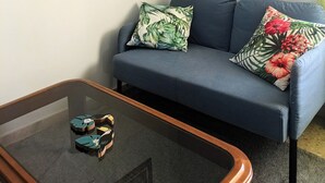 Um quarto com um sofá azul, duas almofadas, uma mesa de centro de vidro com sapatos e duas quadros na parede.