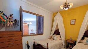 Quarto com duas camas de madeira, penteadeira branca, janela com cortinas, lustre e pequena mesa de cabeceira com abajur.
